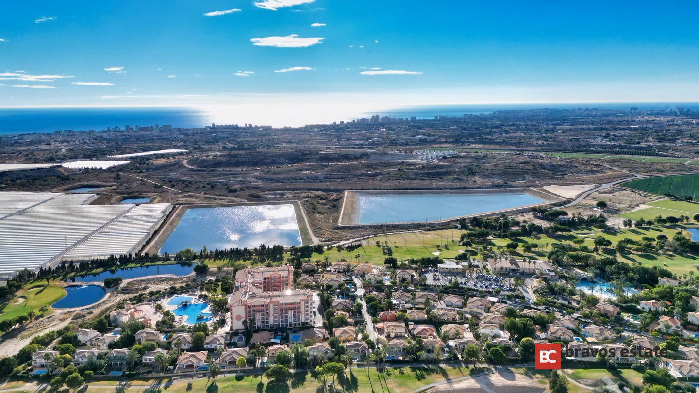 Aerial view of Bonalba Green's golf resort surroundings in Mutxamel, showing the resort hotel, villas, golf fairways, lagoons, and the Mediterranean Sea on the horizon.