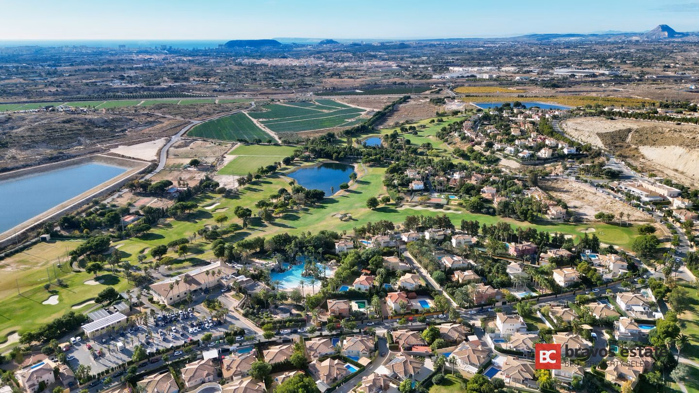Aerial view of Bonalba Green's golf resort setting in Mutxamel, showing the 18-hole course, lakes, residential villas, communal pools, and distant mountains and sea.
