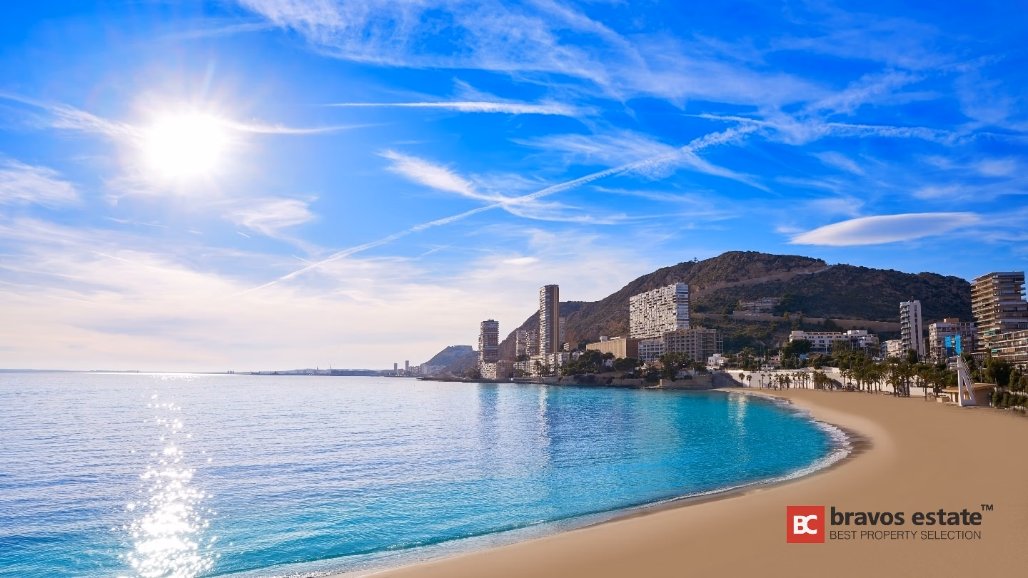 Sandy beach and turquoise Mediterranean Sea near Bonalba Green apartments, with Alicante's coastal skyline and a rocky mountain backdrop under a sunny blue sky.